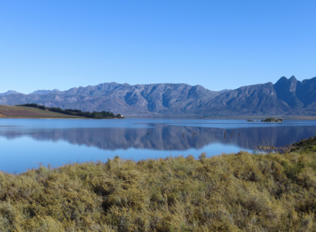 Paysage de lac calme entouré de montagnes et de vignobles près du Cap en Afrique du Sud, ciel bleu dégagé se reflétant dans l’eau, étape nature d’un road trip en 4x4 dans la région du Western Cape