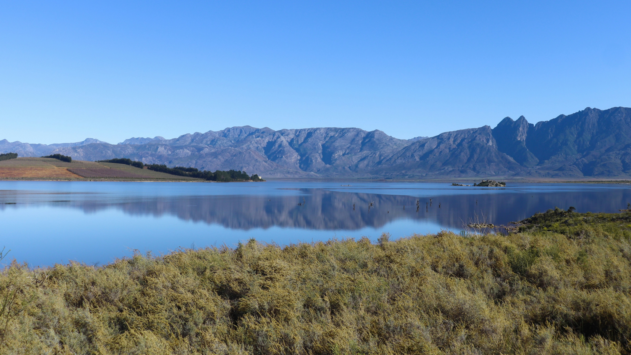 Paysage de lac calme entouré de montagnes et de vignobles près du Cap en Afrique du Sud, ciel bleu dégagé se reflétant dans l’eau, étape nature d’un road trip en 4x4 dans la région du Western Cape
